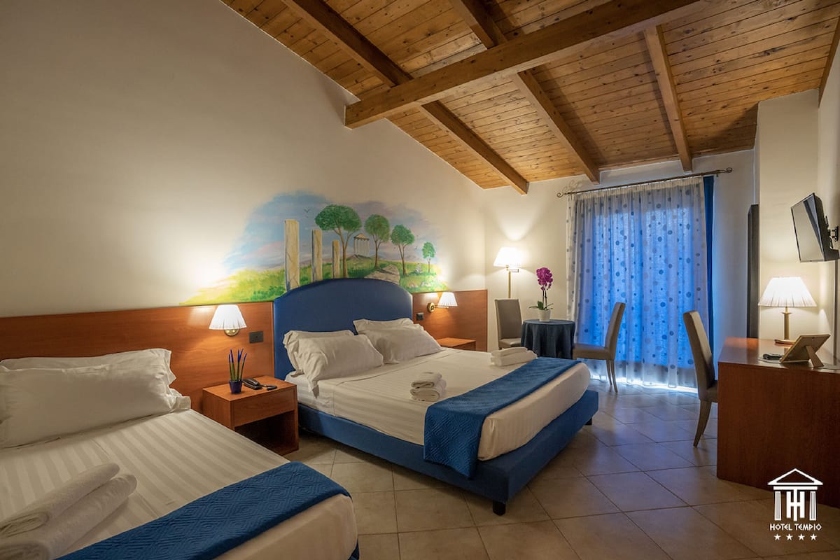 A hotel room with two beds, wooden ceiling beams, a mural on the wall, and a desk with a chair.