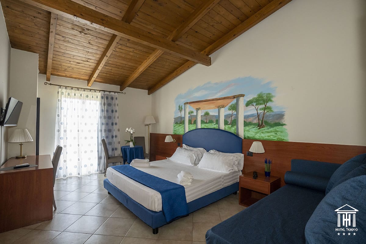 A hotel room with a bed, a desk, a chair, a nightstand, and a painting on the wall.