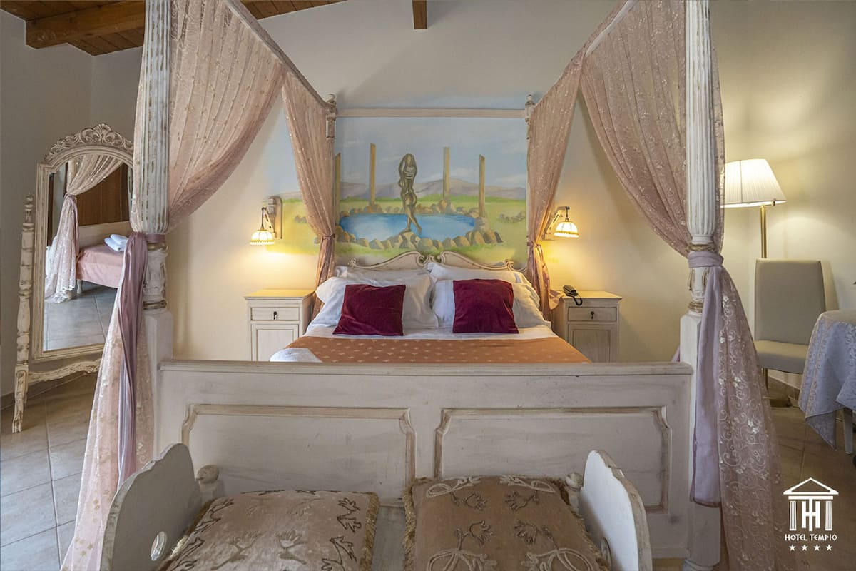A four-poster canopy bed with a painting above it, bedside lamps, and a small table beside the bed.