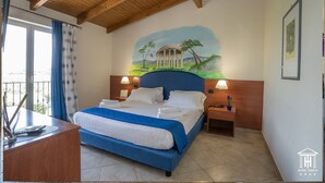 Una camera d'albergo con un letto, due comodini, una scrivania con una sedia, una finestra con tende e un affresco sulla parete.