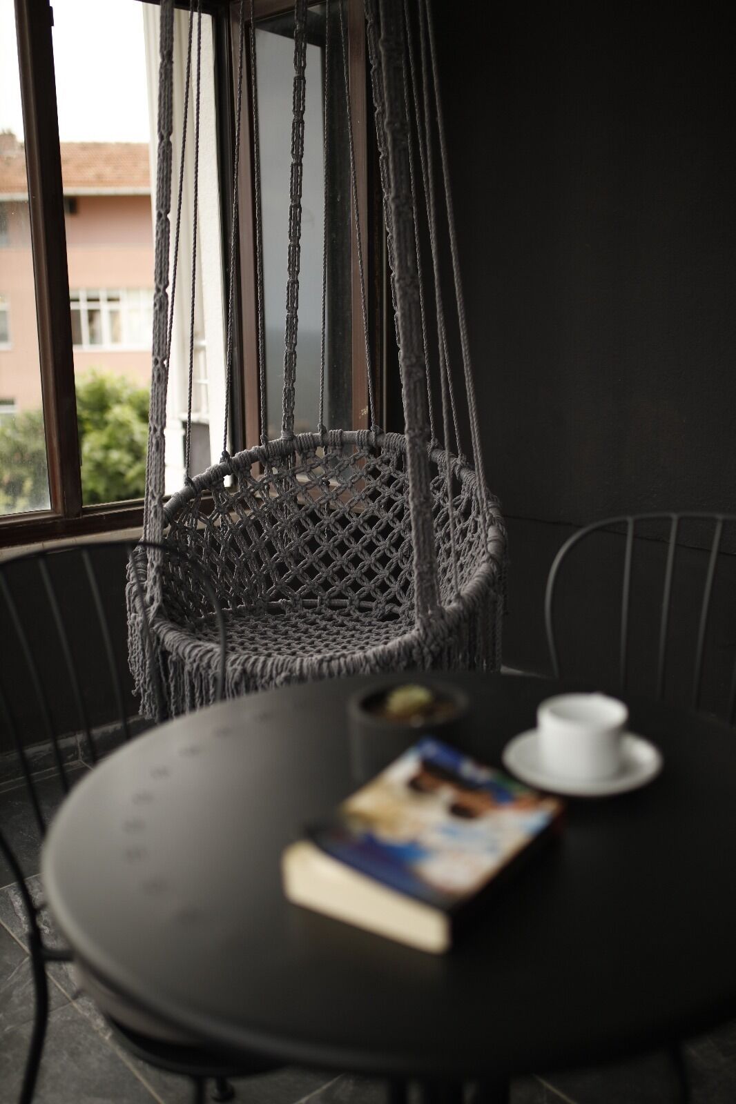 Una habitación con una silla colgante de macramé, una mesa redonda con un libro y una taza, y una ventana con vistas a edificios.