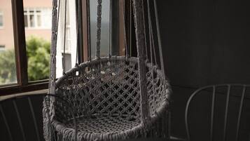 Una habitación con una silla colgante de macramé, una mesa redonda con un libro y una taza, y una ventana con vistas a edificios.