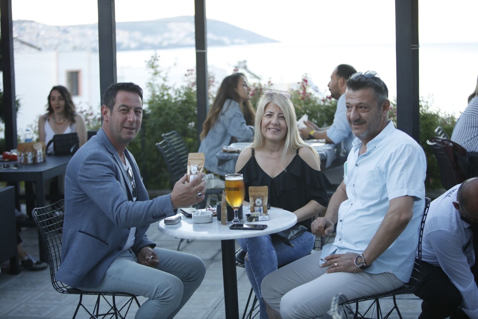 Un grupo de personas sentadas en una mesa al aire libre, con vista al mar y las montañas de fondo.
