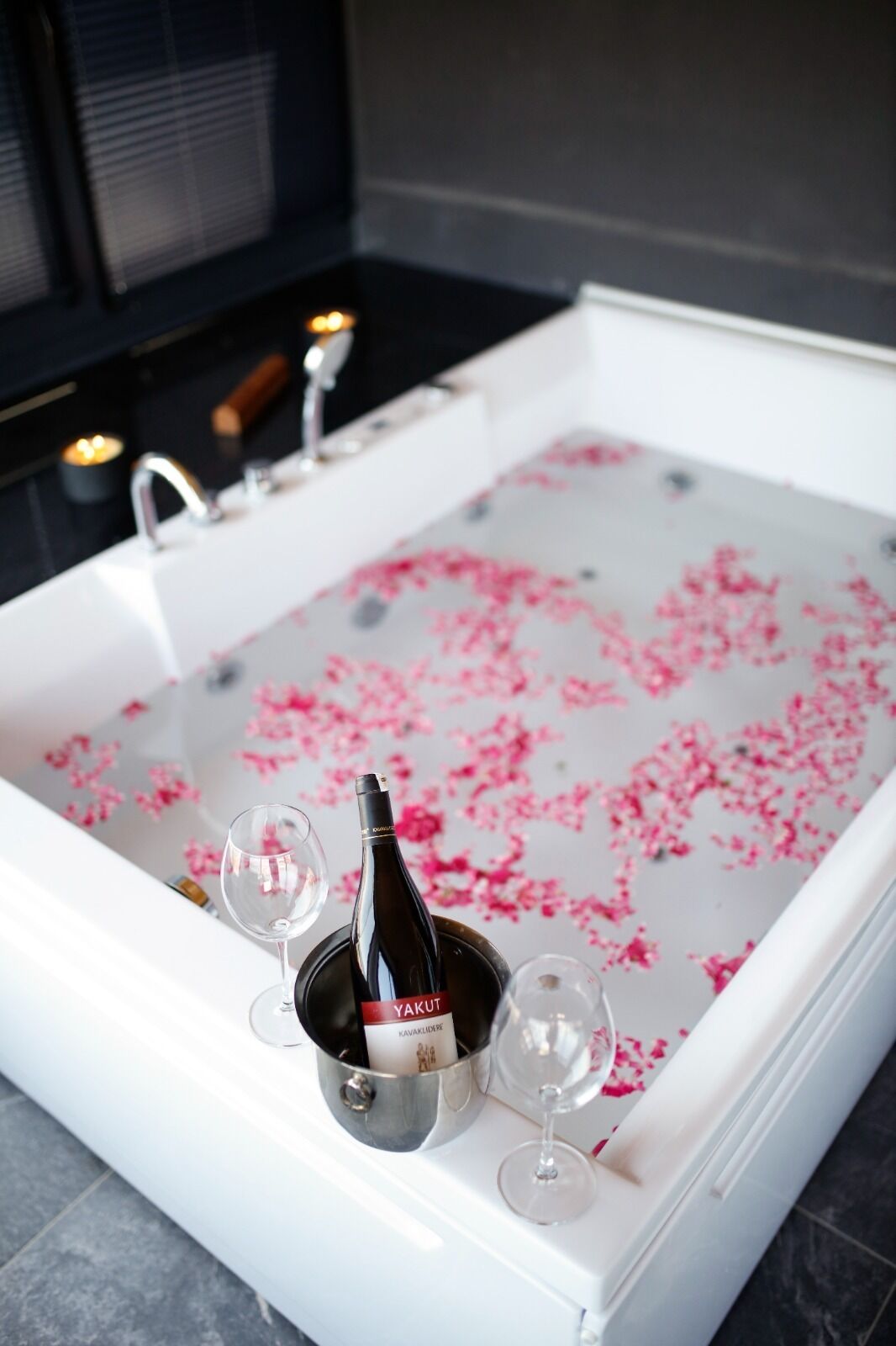A bathtub filled with water and pink flower petals, a bottle of wine, and two wine glasses.