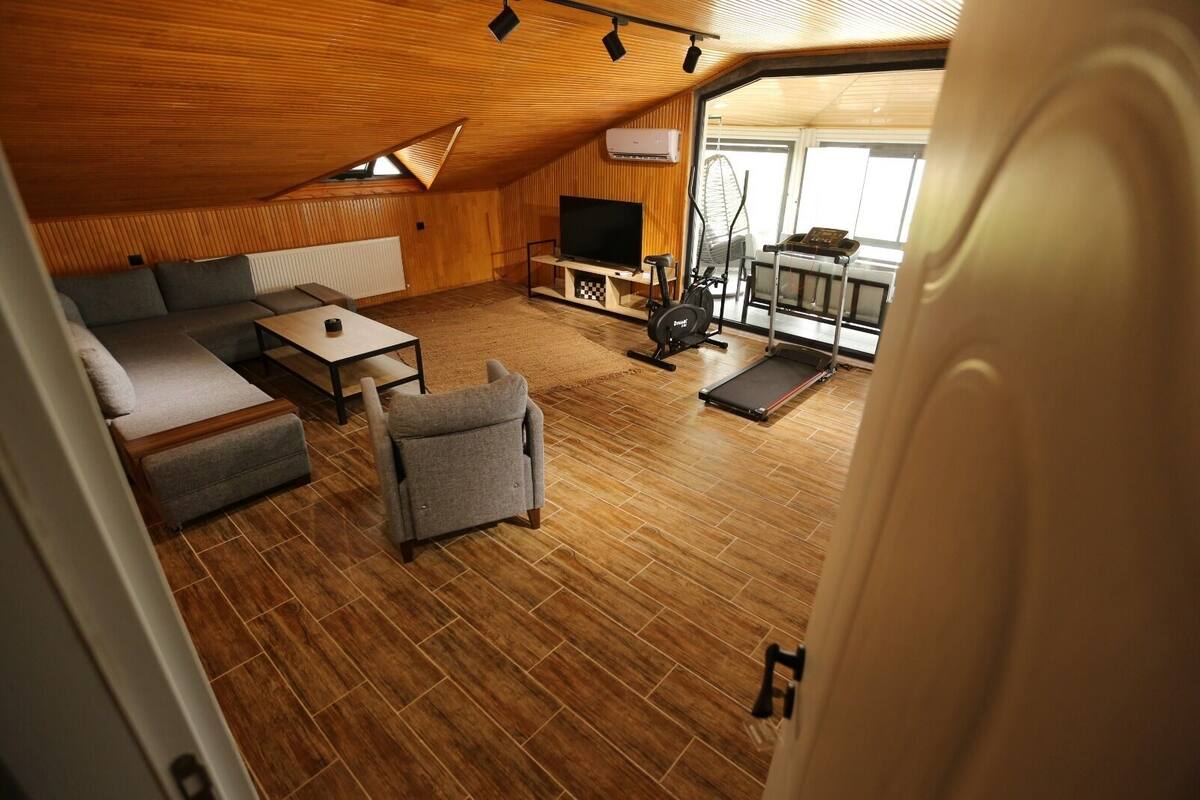 Un espacio habitable compacto con sofá, reposapiés, televisor, bicicleta estática y una pequeña mesa.
