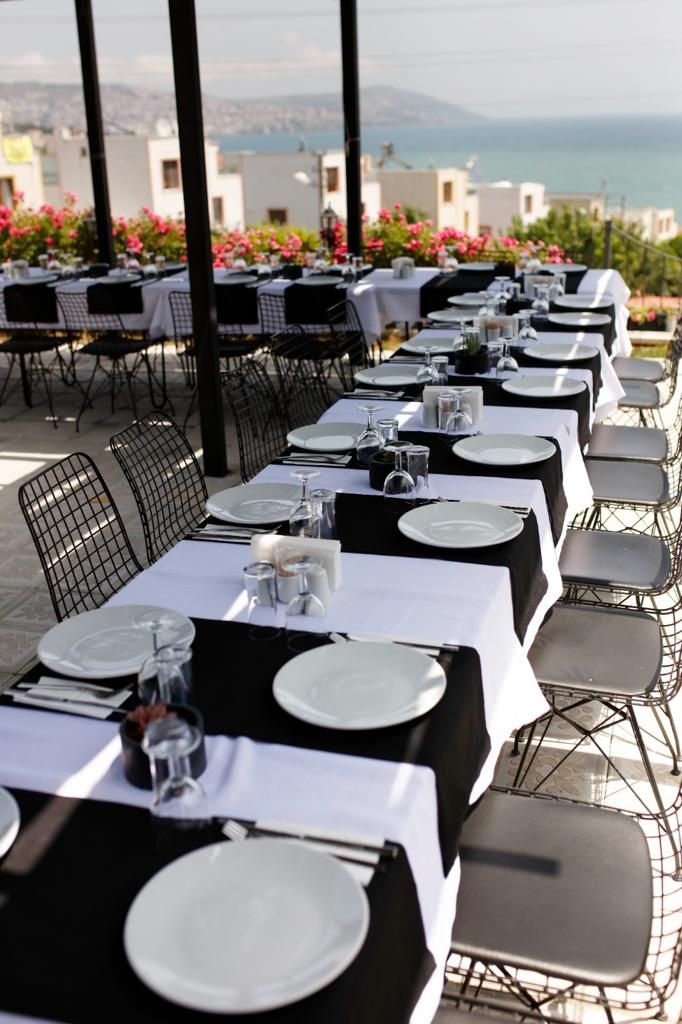 A row of tables set for dining with chairs, a view of the sea, and buildings in the background.