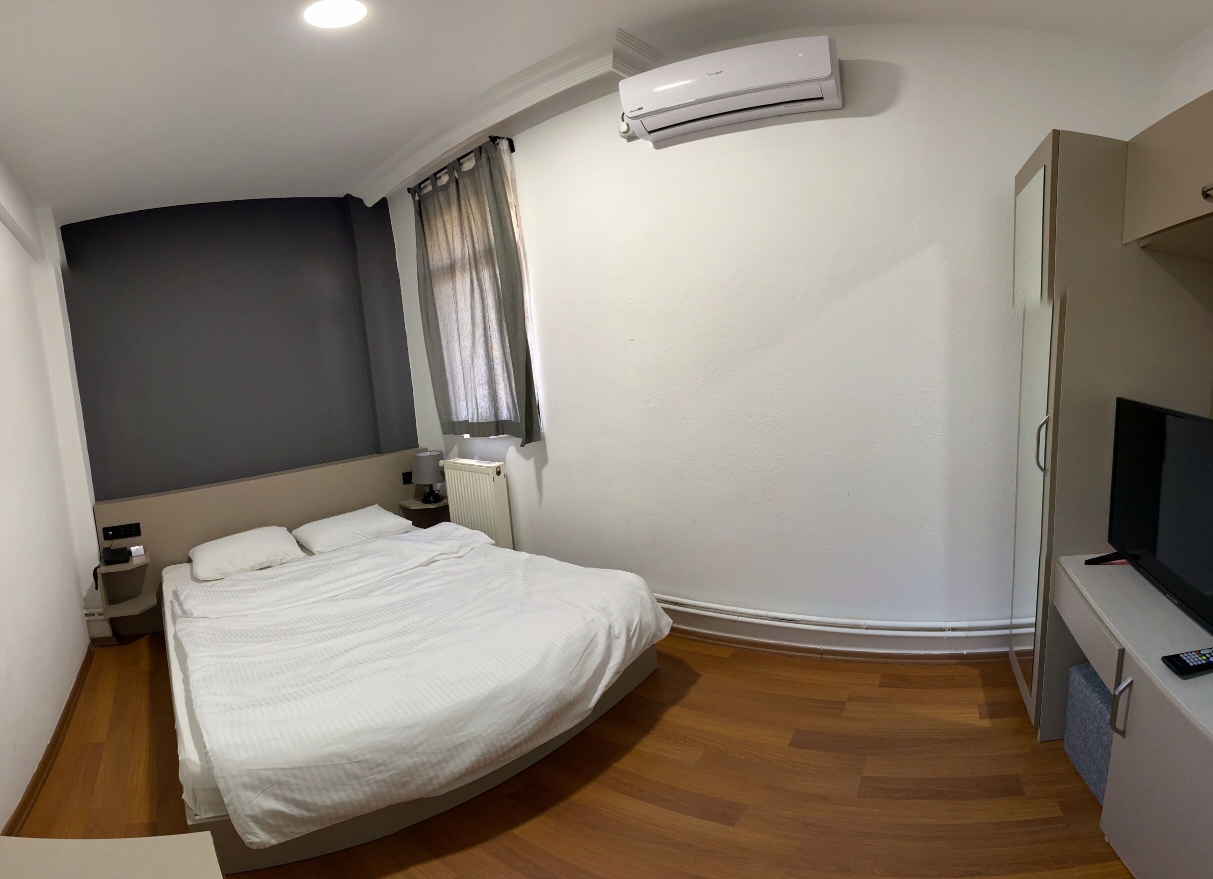 Une chambre d’hôtel comprenant un lit, une télévision, un climatiseur, une fenêtre avec des rideaux, une table de chevet et un radiateur.