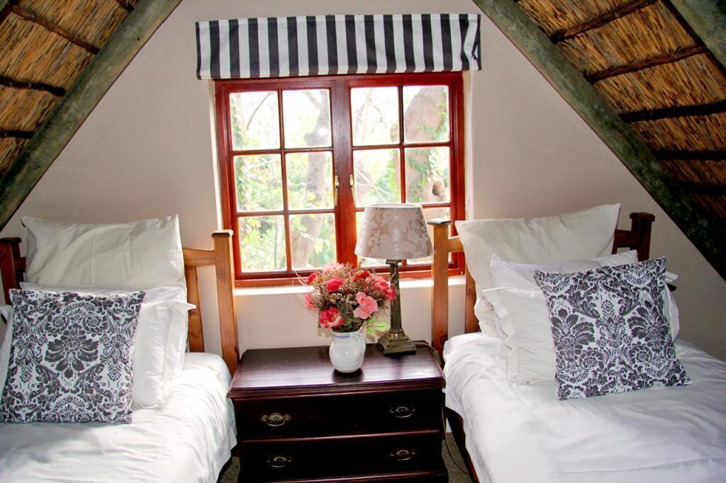 Un dormitorio con dos camas, una ventana adornada con un valance a rayas, una mesita de noche con una lámpara y flores, y techo de madera.