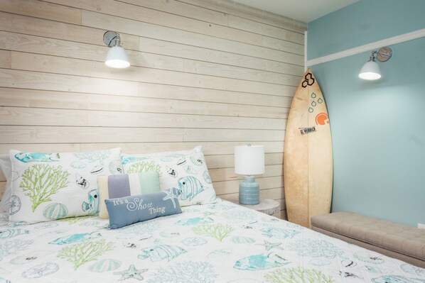 A bedroom with a wooden headboard, a bed with a sea-themed duvet and pillows, a surfboard, and a nightstand with a lamp.