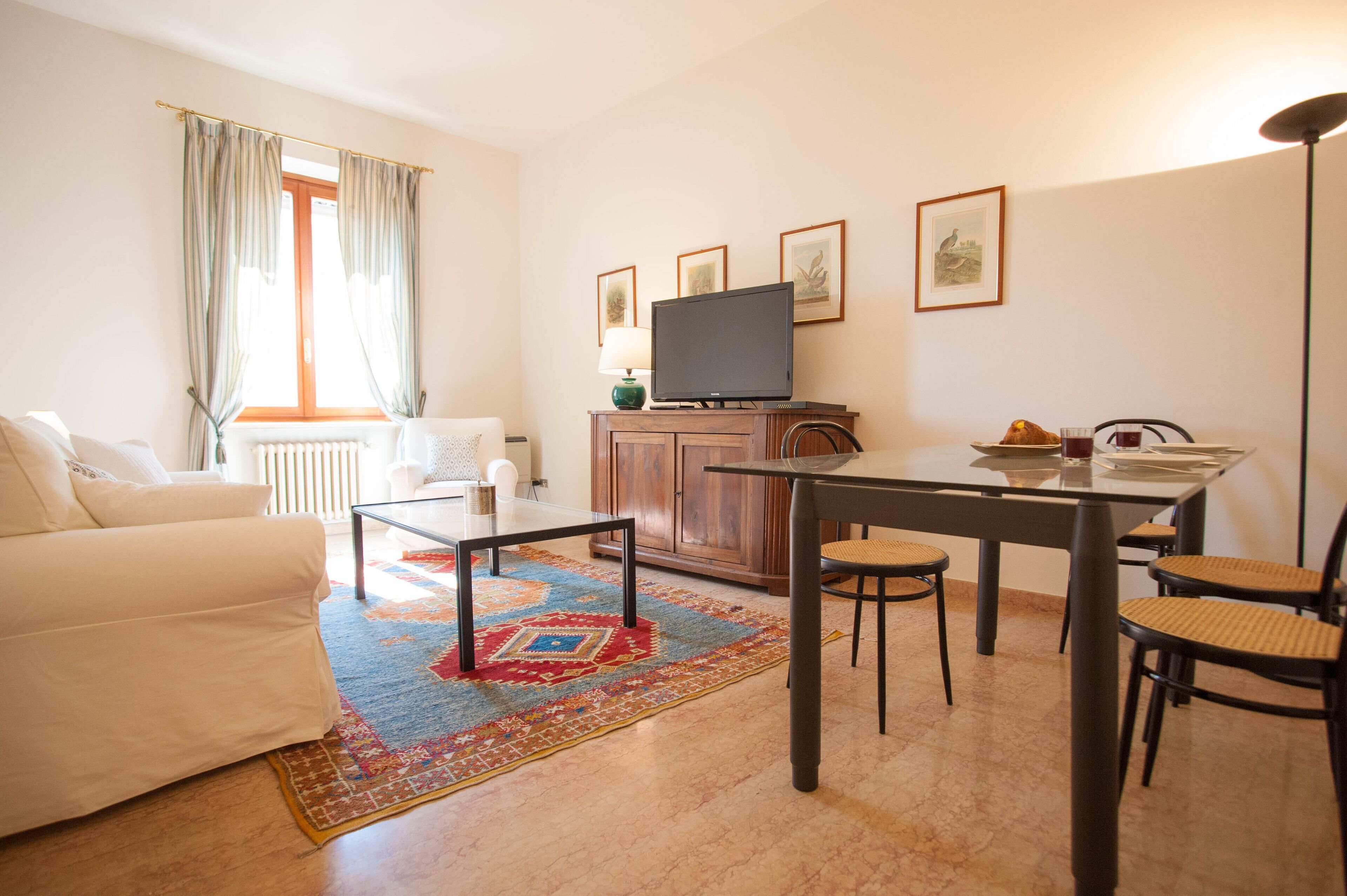 A living room with a sofa, a coffee table, a TV on a cabinet, a dining table with chairs, and a patterned rug.