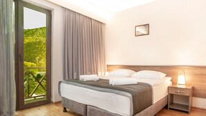 Habitación de hotel con cama, mesita de noche, lámpara, ventana con cortinas y vista a zonas verdes.