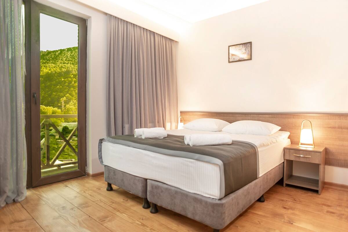 A hotel room with a bed, a nightstand, a lamp, a window with curtains, and a view of greenery outside.