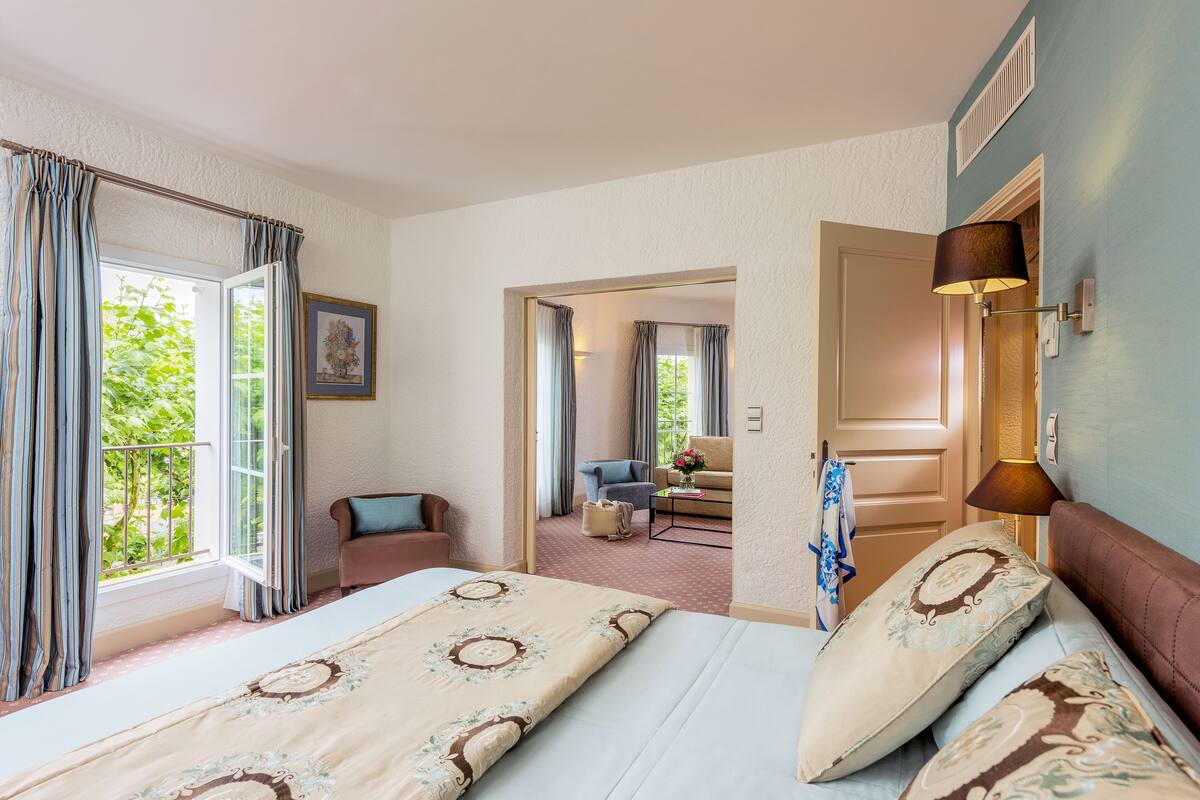 Een hotelkamer met een groot bed, een zithoek met een bank en fauteuil, een kleine tafel met een bloevas, en uitzicht op groen.
