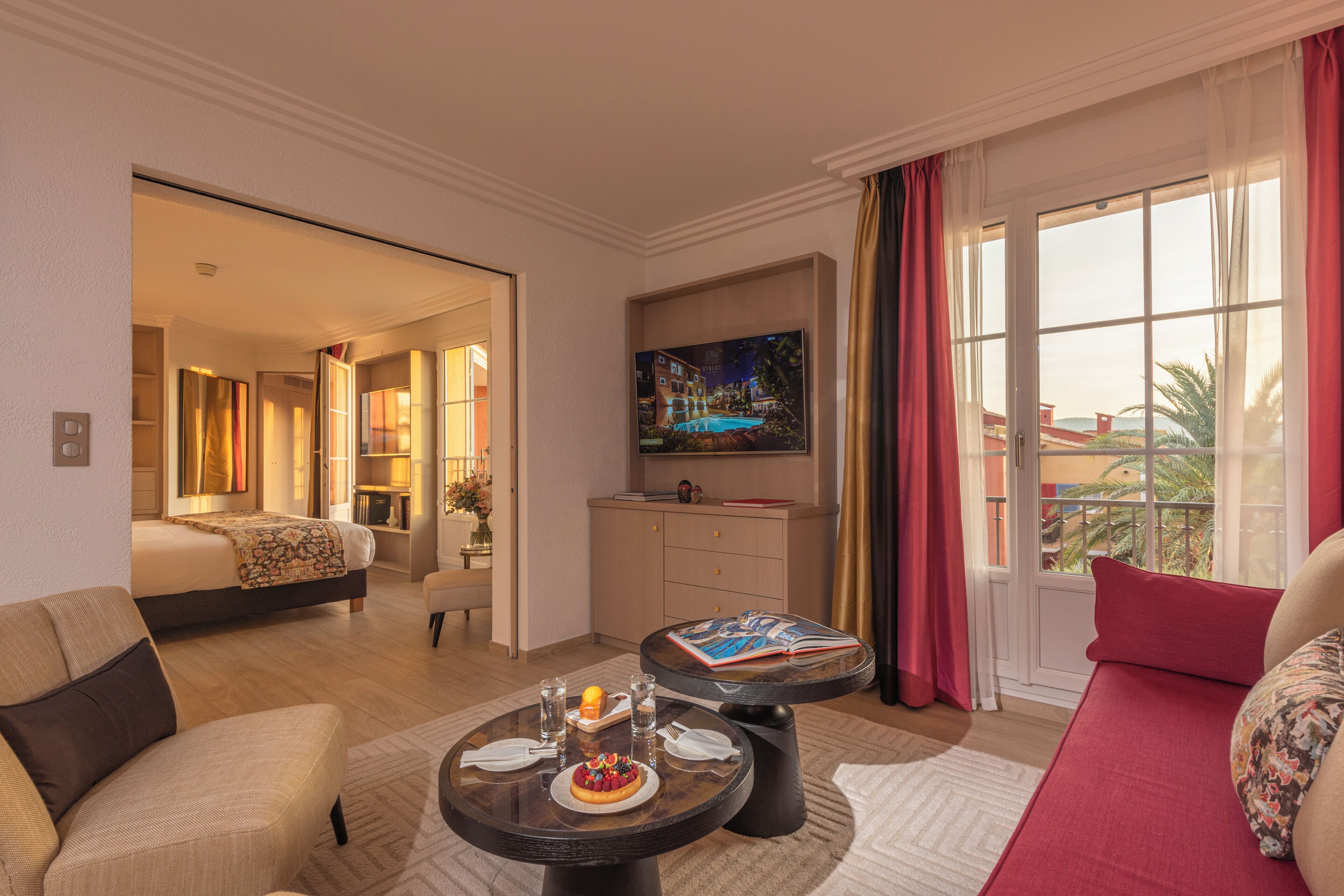 Habitación de hotel con cama, zona de comedor con mesa preparada para dos personas, televisor montado en la pared y un gran ventanal con cortinas.