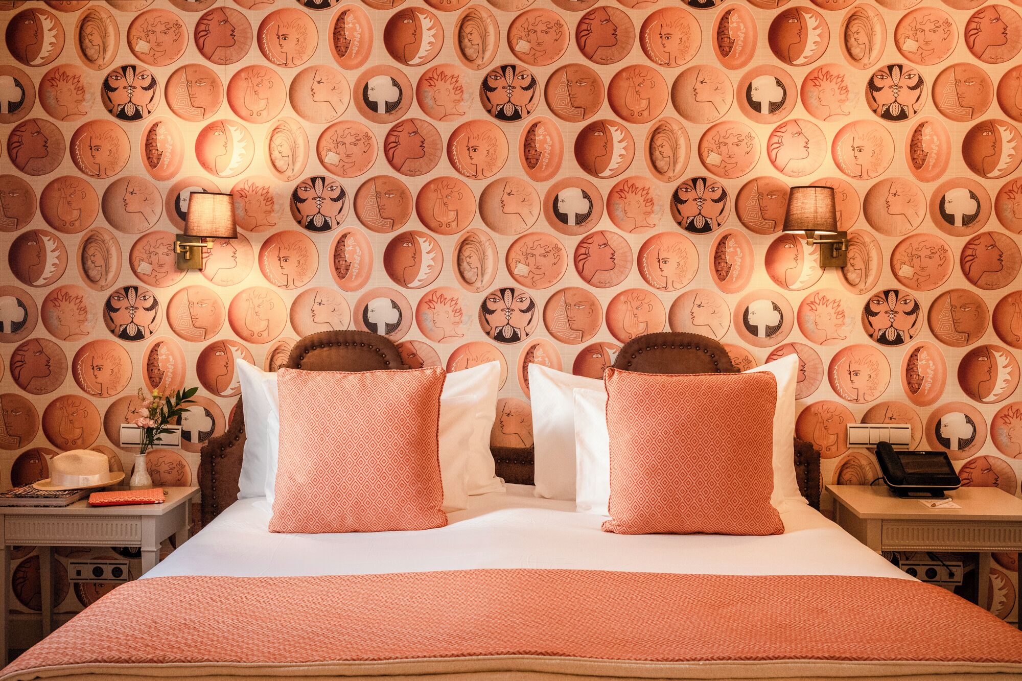A hotel room with a patterned wall, a bed with two pillows, a bedside table with a lamp, and a nightstand with a phone.