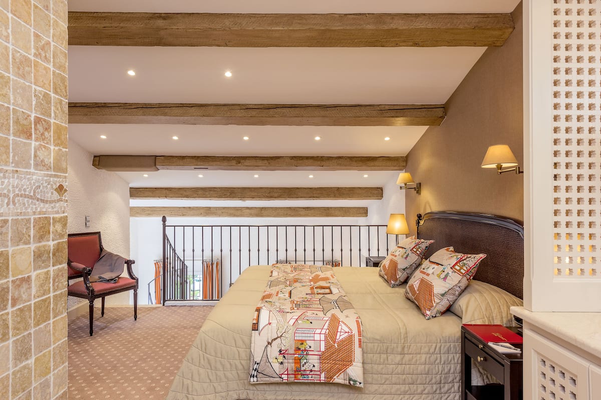 Een slaapkamer met een groot bed, houten balken in het plafond, een bankje en uitzicht op een balkon.