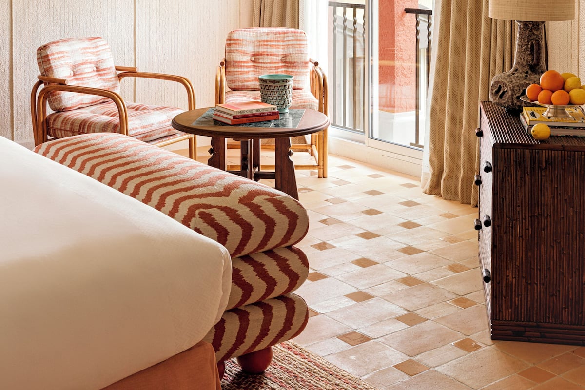 Hotelkamer met een bed, twee fauteuils, een tafeltje met boeken en een mok, een lamp en een fruitmand.