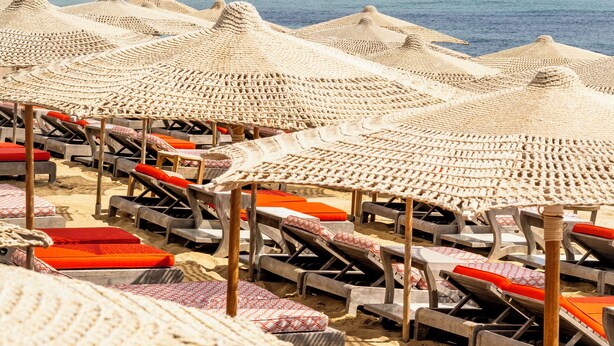 Een strand met rijen ligbedden en parasols, een helder uitzicht op de zee, en een verafgelegen groene heuvel.
