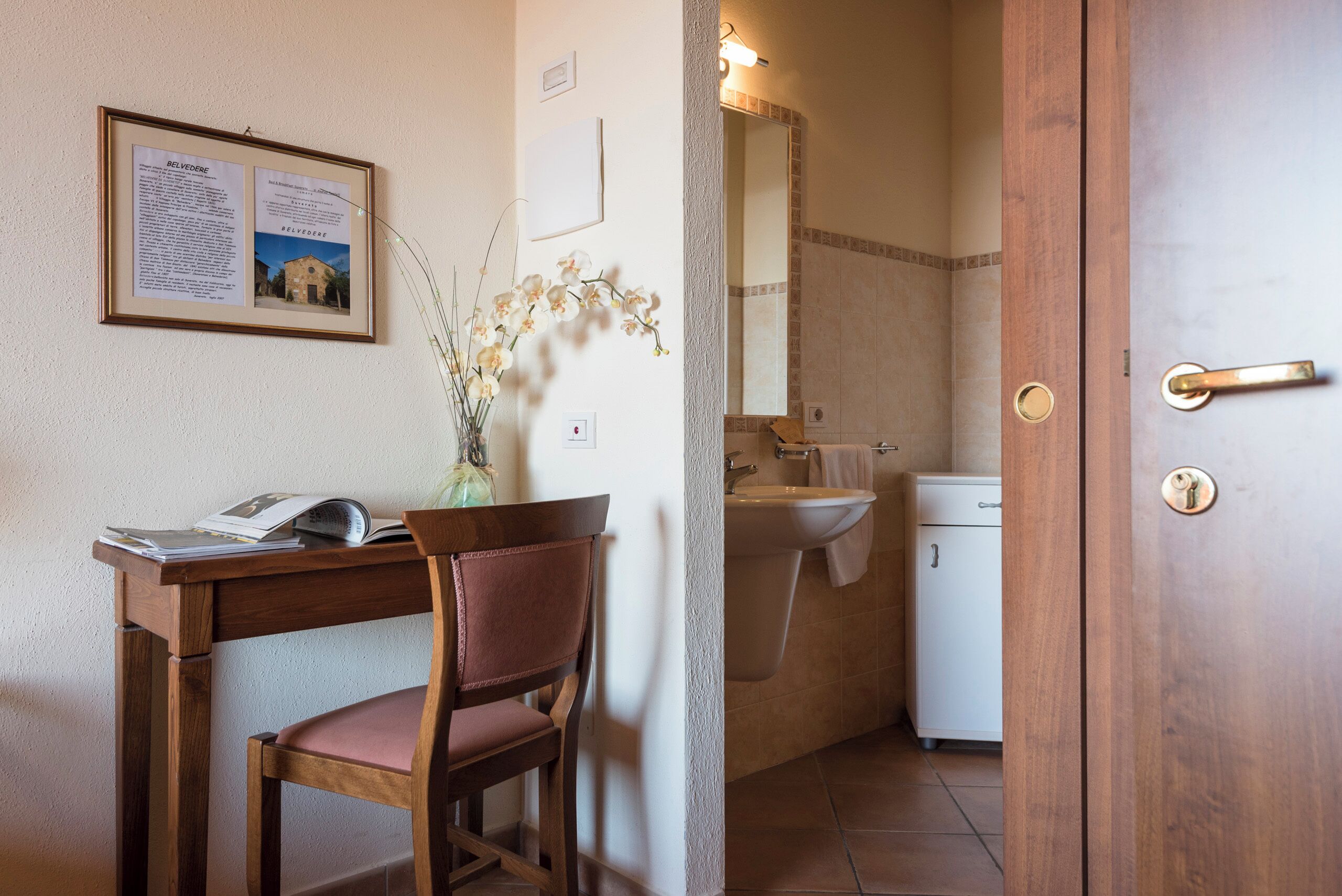 Un baño con lavamanos, espejo y un mueble pequeño. Hay un cartel informativo enmarcado en la pared y una silla con un libro.
