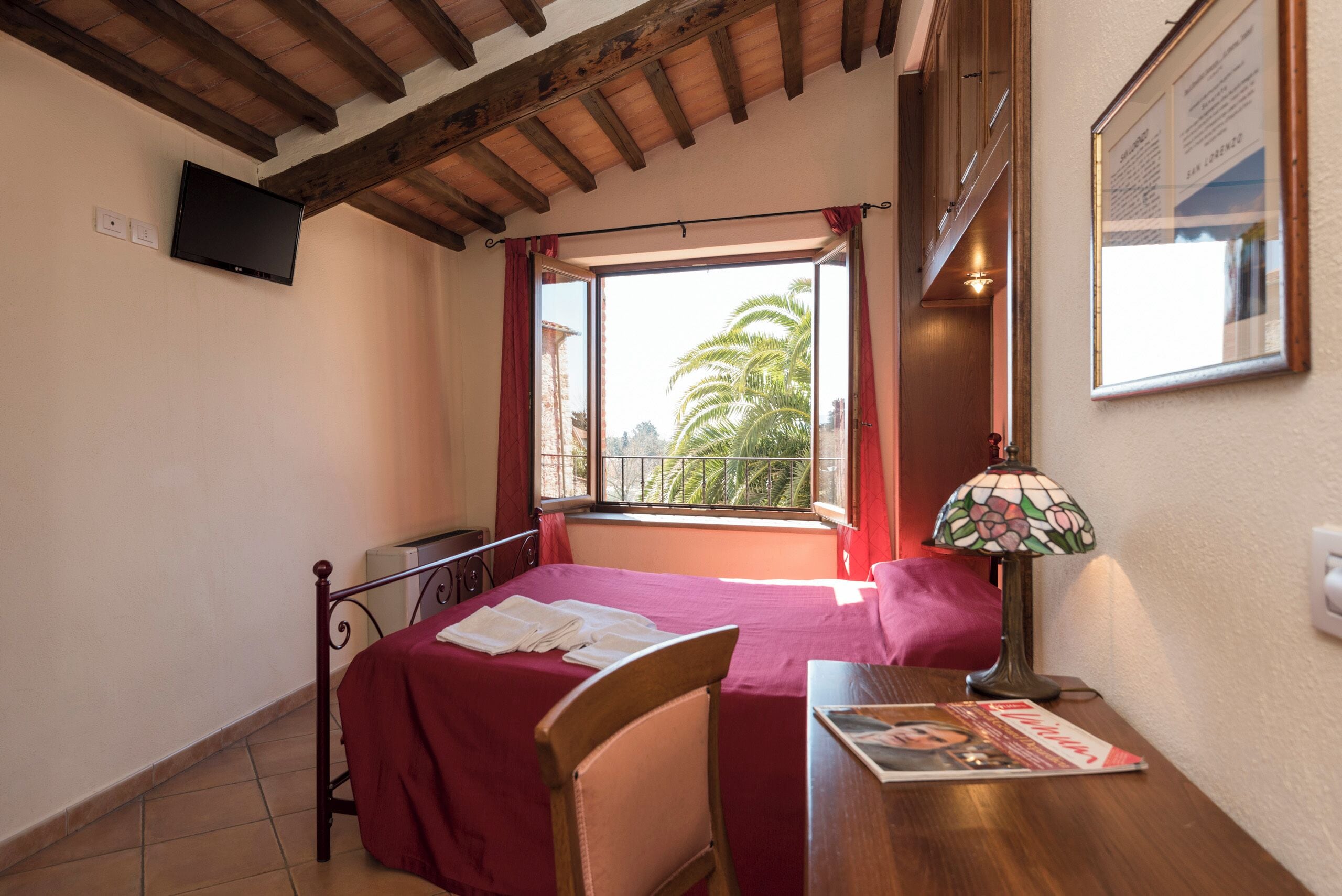 A bedroom with a bed, a desk, a chair, a lamp, and a window with a view of palm trees.