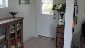 Een gang met een witte deur, een houten kast met decoratieve kussens, een vaas met bloemen en een boekenkast vol boeken.