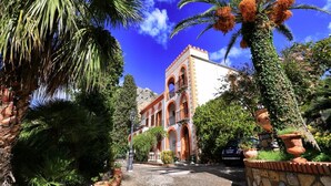 A building with arched windows and a stone facade, surrounded by lush greenery and palm trees. - Solemar Sicilia - Villa Caterina (Cefalù)