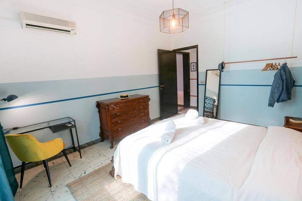 A bedroom with a bed, a wooden dresser, a desk with a chair, an air conditioner, and a hanging light fixture.