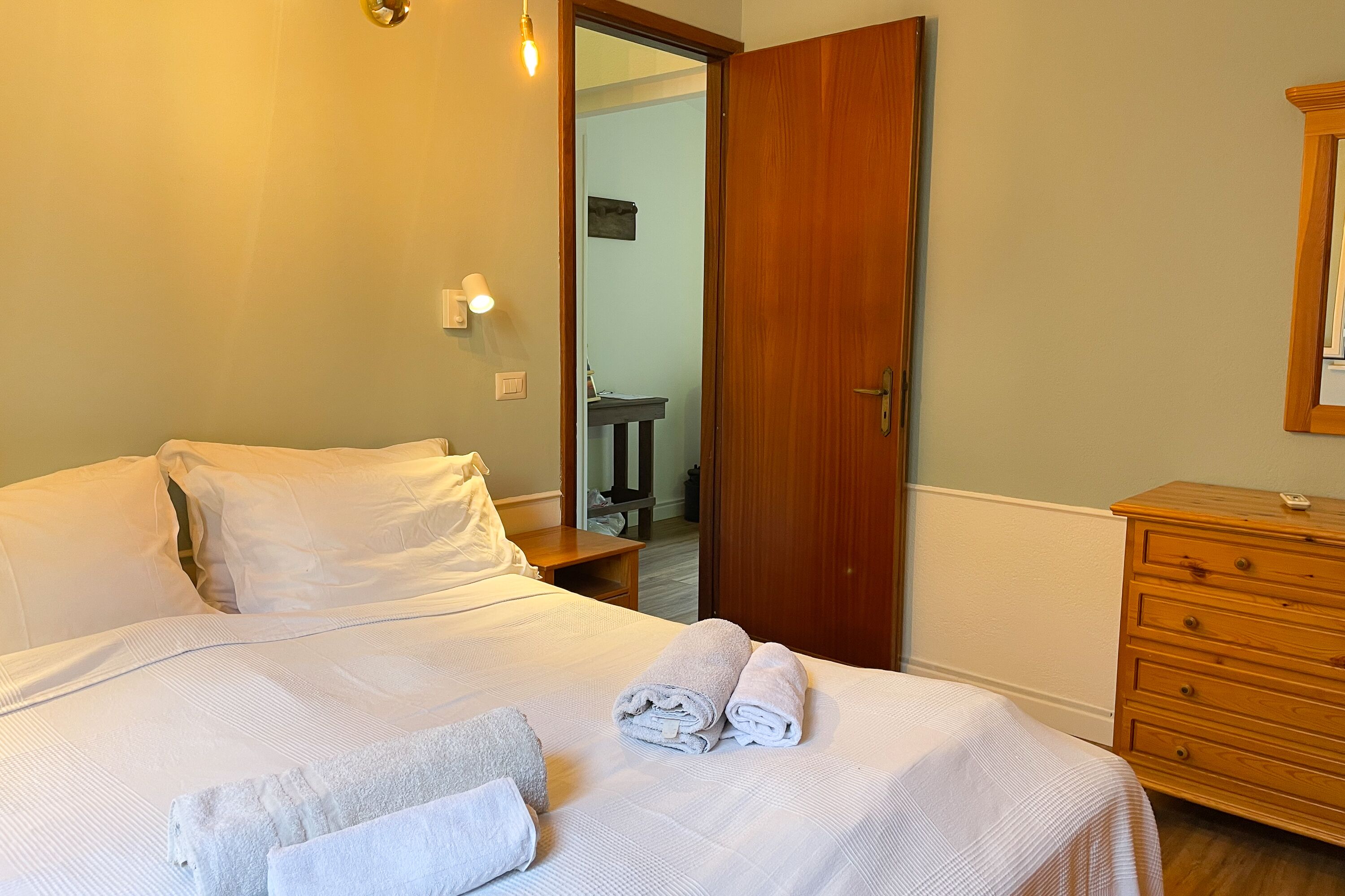A neatly made bed with white linens and towels, a wooden dresser, and a door leading to another room.