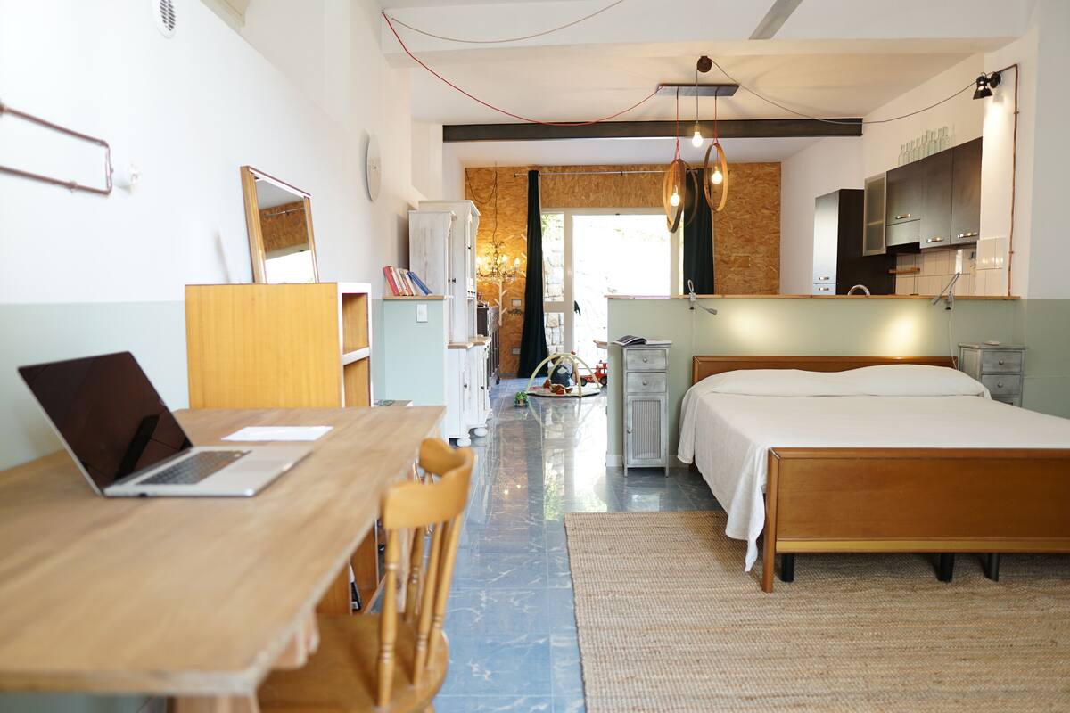 A modern hotel room with a wooden desk, laptop, and chair, a large bed, and a kitchenette area.