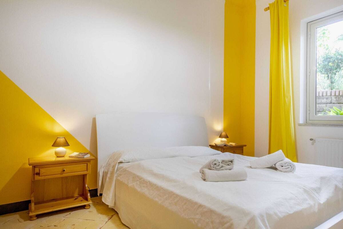 A bedroom with a bed, two bedside tables, and a window with yellow curtains.