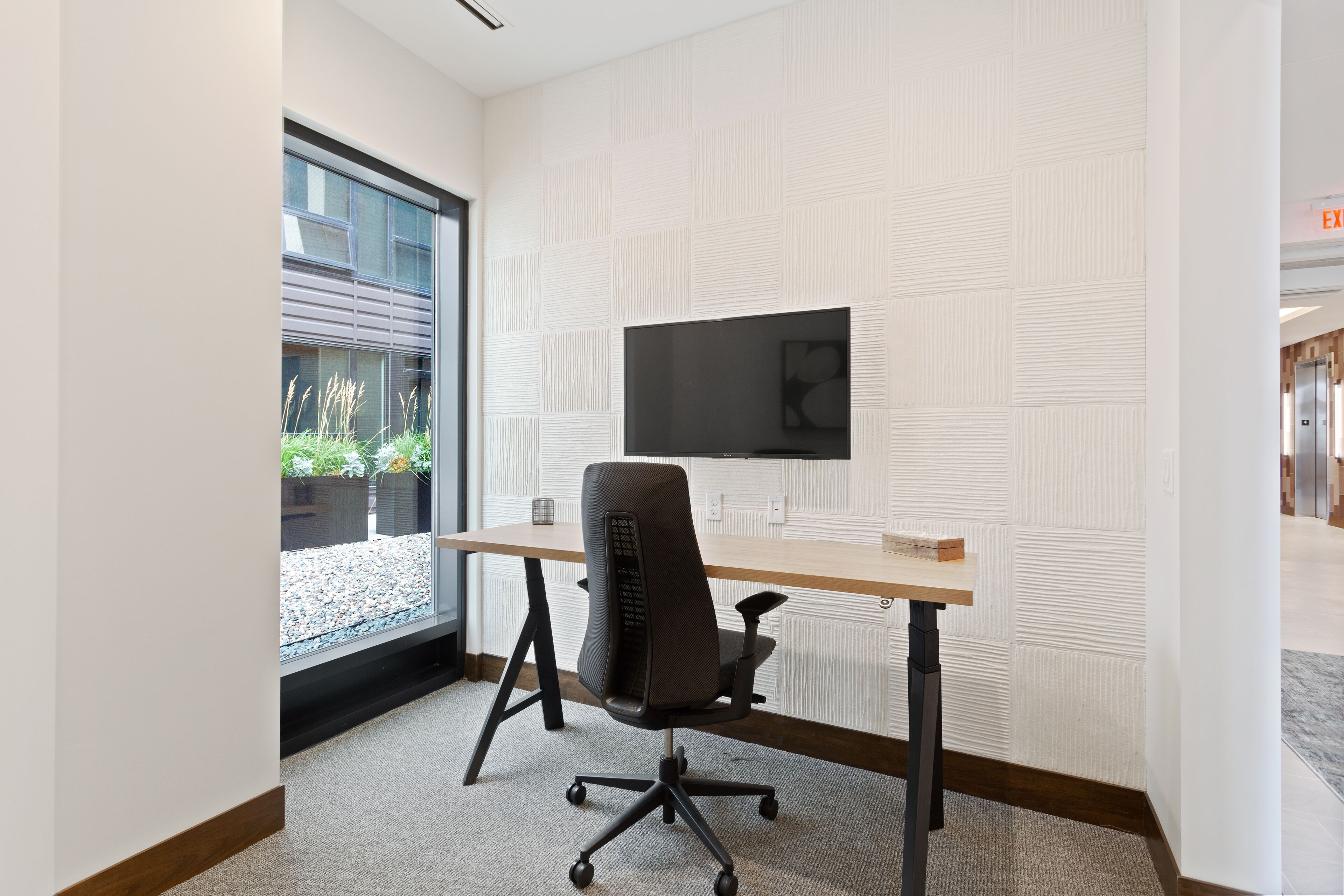 A modern office space with a large desk, a black office chair, a mounted flat-screen TV, and a window with a view of plants and a building.
