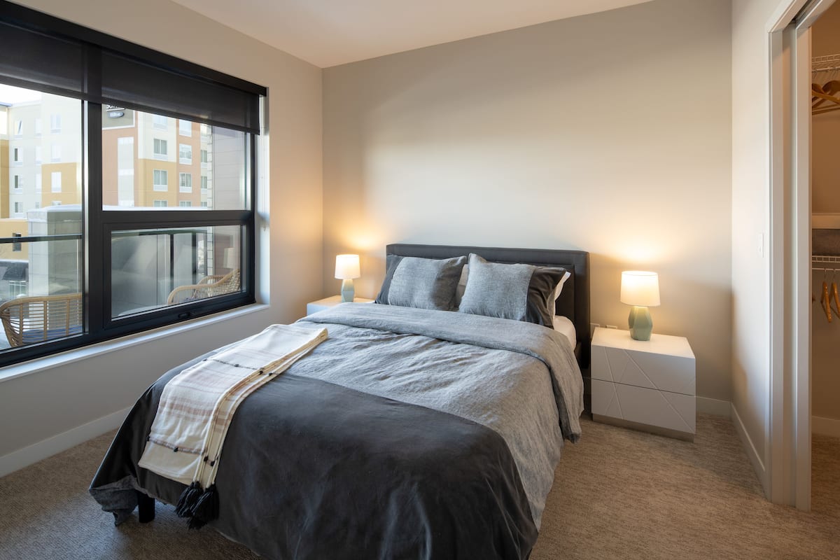 A bedroom with a large window, a bed with a gray and white duvet, two bedside tables with lamps, and a view of buildings outside.