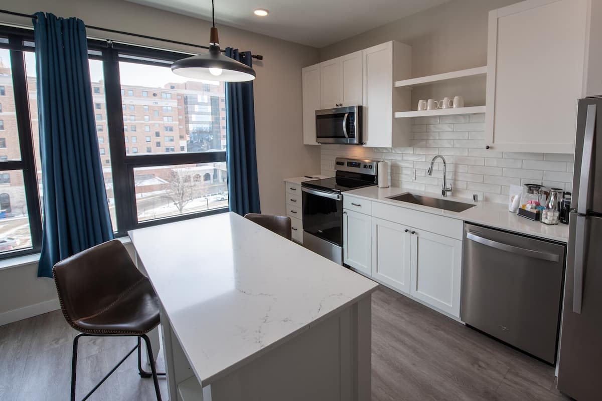 A modern kitchen with white cabinets, stainless steel appliances, a marble countertop, and a view of the city through large windows.