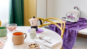 Ein Zimmer mit Esstisch, gedecktem Tisch, Stuhl, Bett mit violetter Decke und weißem Rucksack mit Zeichentrickfiguren.