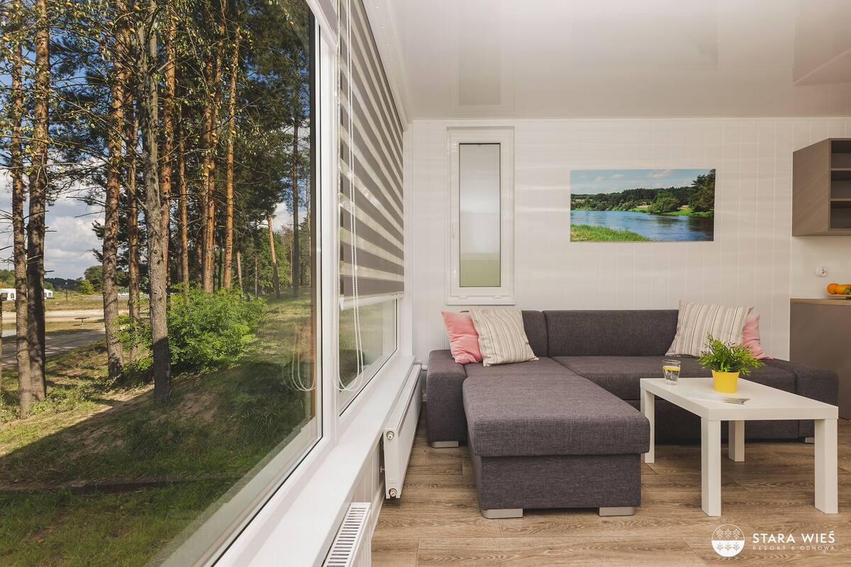 A modern living room with a grey sectional sofa, a white coffee table, and a view of a forest through large windows.