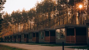 A row of cabins with large windows and wooden railings, set against a backdrop of tall trees and a setting sun. - Resort Stara Wieś (Patnów)