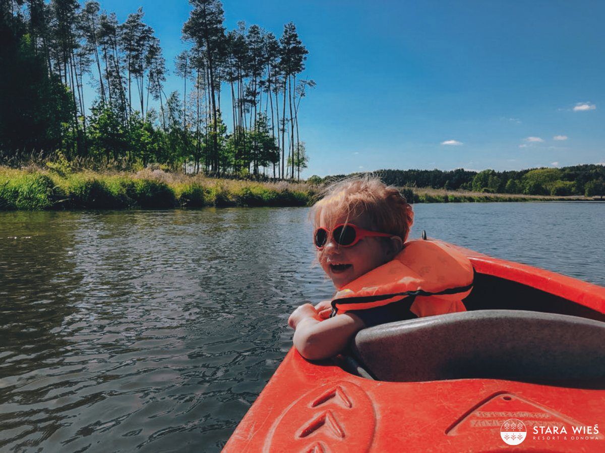 A child in a red kayak on a calm river, surrounded by tall trees and a clear sky.