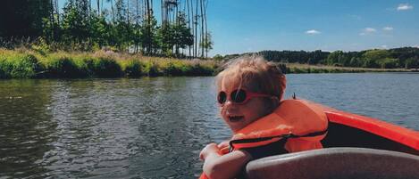 Un niño en un kayak rojo en un río tranquilo, rodeado de altos árboles y un cielo despejado.