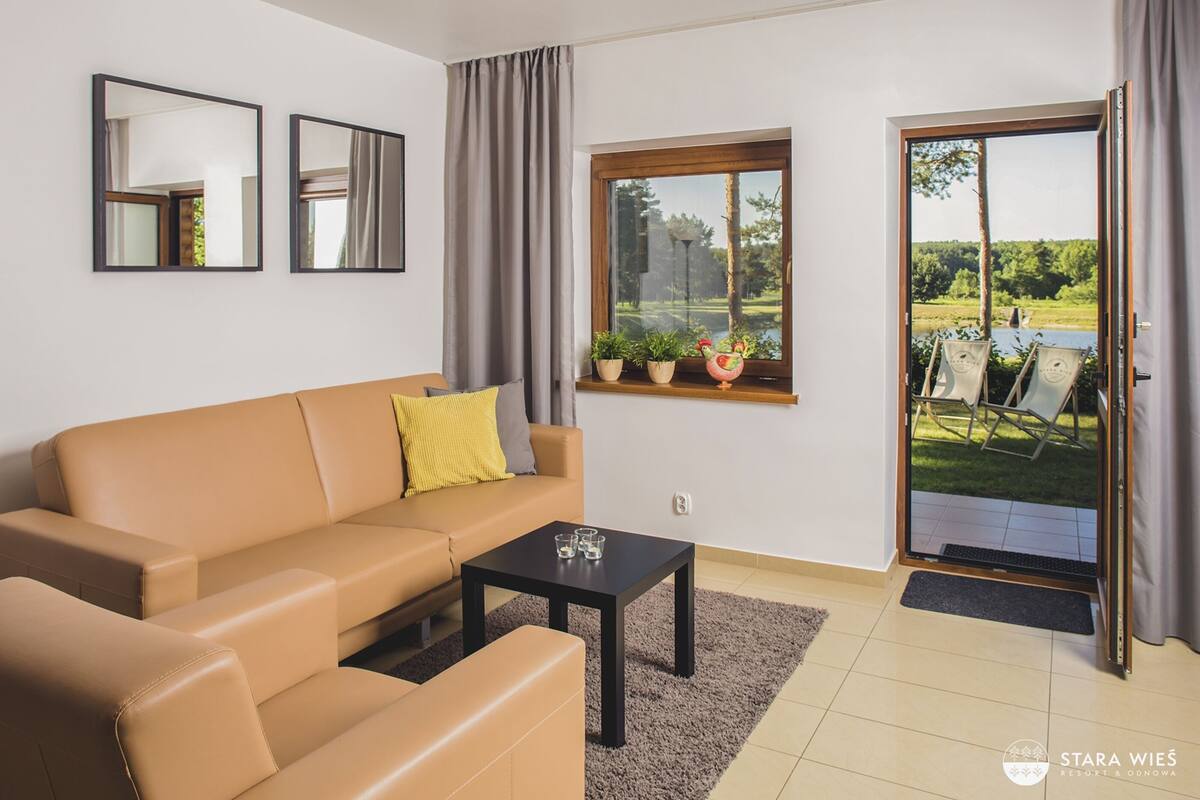 A modern living room with a beige sofa, a black coffee table, and a window with potted plants.