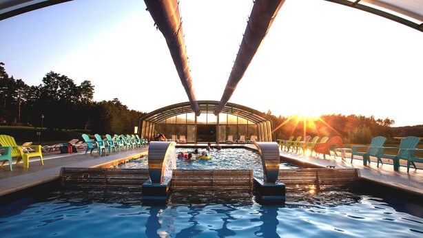 A swimming pool with a glass ceiling, surrounded by lounge chairs and a sunlit landscape. - Resort Stara Wieś (Patnów)