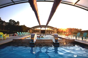 Una piscina con techo de cristal, rodeada de sillones de descanso y un paisaje bañado por el sol.