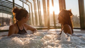 Two people in a hot tub with a view of a modern building and the sun setting in the background. - Resort Stara Wieś (Patnów)