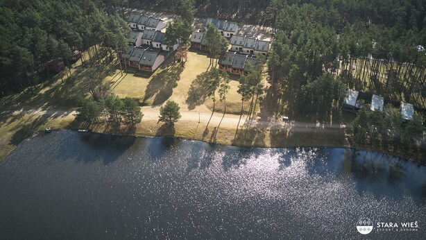 An aerial view of a lakeside resort with multiple buildings, a sandy beach, and dense forest surrounding the area. - Resort Stara Wieś (Patnów)