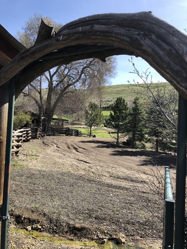 An outdoor area with a wooden archway, a dirt path, and a fenced area with trees and a hill in the background.