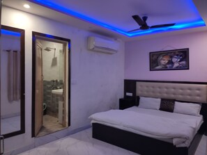 A bedroom with a bed, a ceiling fan, an air conditioning unit, and a bathroom visible through a glass door. - Hotel Hare Rama Deluxe (New Delhi)