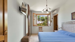 A bedroom with a bed, a window with a view of palm trees, a chair, a bedside table, and a wall-mounted TV. - Villa Mila by Perle di Puglia (Torre Vado)