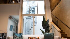 A modern living room with a white sofa, geometric patterned cushions, a large plant, and a view of a spiral staircase through a large window. - Luxury apartment with a private rooftop deck located in the heart of downtown JC (Johnson City)