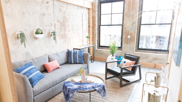 A living room with a grey sofa, a small round coffee table, a chair, and a large window with a view of buildings. - Luxury apartment with a private rooftop deck located in the heart of downtown JC (Johnson City)