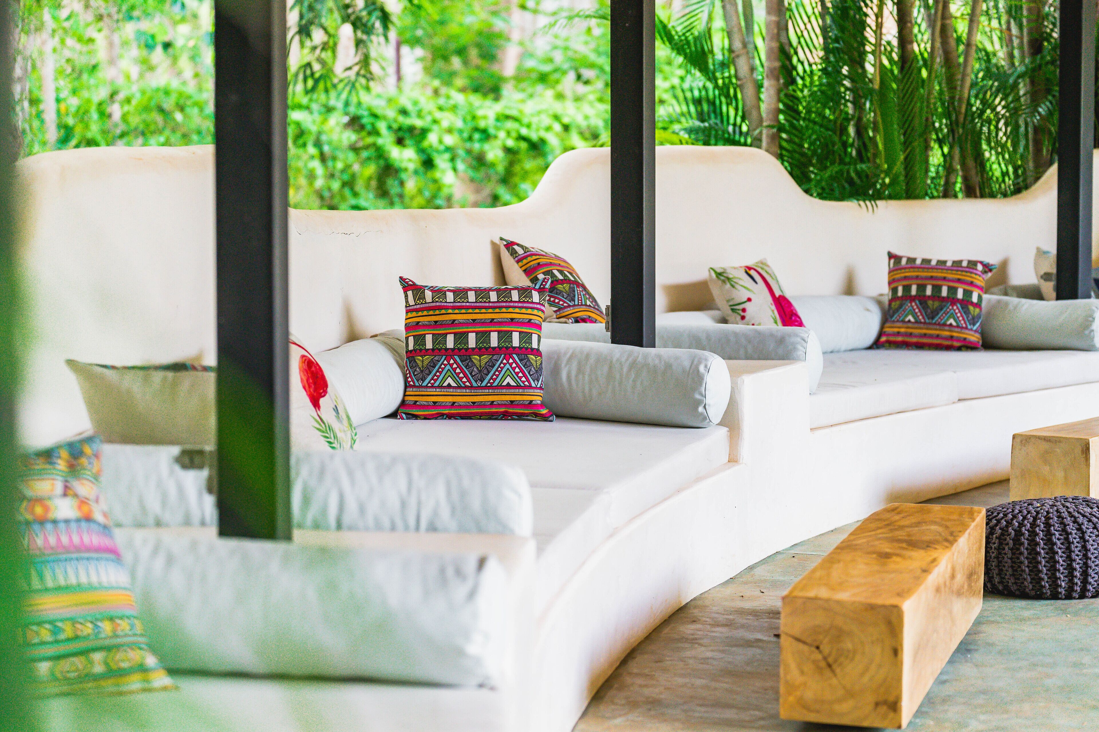 Un sofá blanco curvo con almohadones coloridos, un banco de madera y vistas a abundante vegetación.