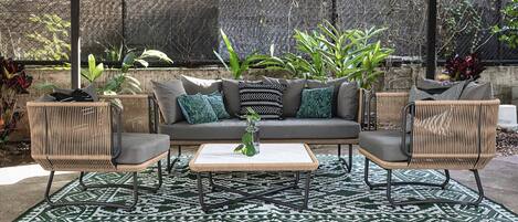 An outdoor seating area with a sofa, two armchairs, and a coffee table, surrounded by a patterned rug and greenery.